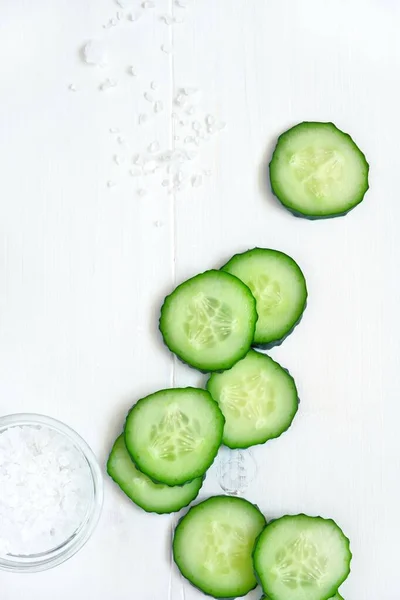 Sliced cucumber slices, salt, pepper, spices in glass bowls on a white wooden table. Freshness concept, vegetarianism, salad ingredients. Top view, flat lay, copy space for text.