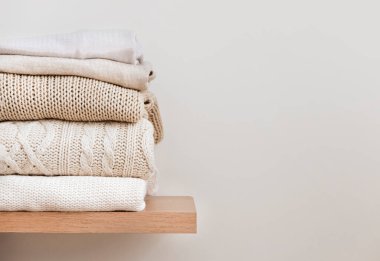 Bir yığın düzgün katlanmış kalın örgü, ahşap bir rafta yün ve bej bir arka plan. Kapsül dolabı, giysi deposu, minimalizm, örgü dokuma, düzen, rahatlık. Metin için kopyalama alanı olan pankart.
