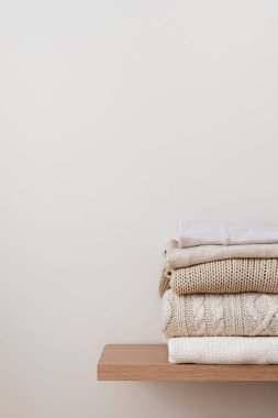 Bir yığın düzgün katlanmış kalın örgü, ahşap bir rafta yün ve bej bir arka plan. Kapsül dolabı, giysi deposu, minimalizm, örgü dokuma, düzen, rahatlık. Dikey, boşluğu kopyala.