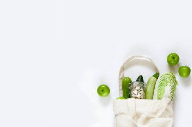 Yeşil elmalı tekrar kullanılabilir keten torbası, cam kavanozda kabak çekirdeği, beyaz arka planda taze sebzeler. Sıfır atık alışverişi, sağlıklı beslenme, sebze yemeği kavramı. Düz konum, üst görünüm, kopyalama alanı.
