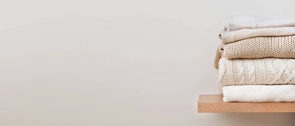 Bir yığın düzgün katlanmış kalın örgü, ahşap bir rafta yün ve bej bir arka plan. Kapsül dolabı, giysi deposu, minimalizm, örgü dokuma, düzen, rahatlık. Metin için kopyalama alanı olan pankart.