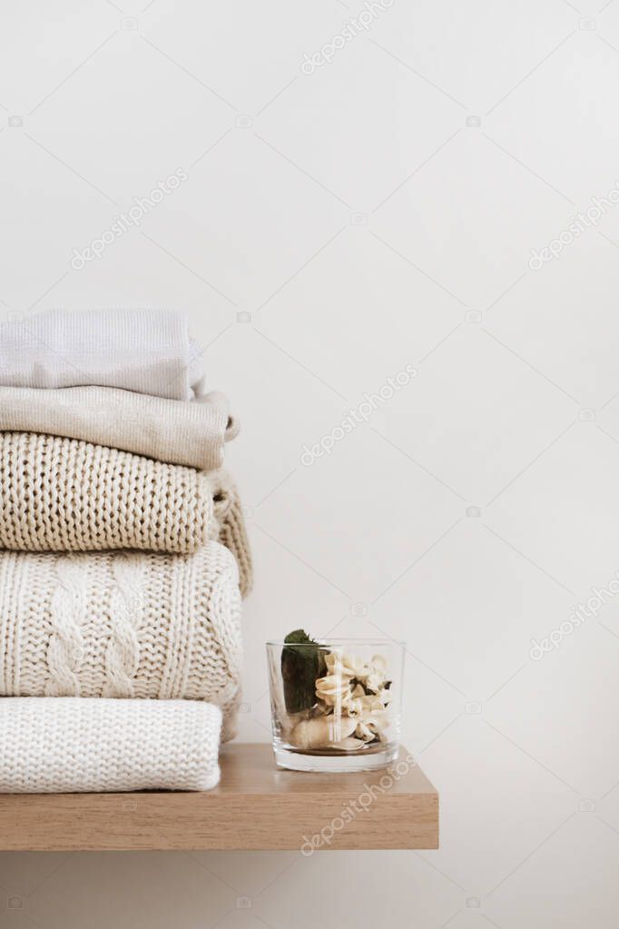 A stack of neatly folded warm knitwear, wool on a wooden shelf and a beige background. Capsule wardrobe, clothes storage, minimalism, knitted texture, order, comfort. Vertical, copy space.