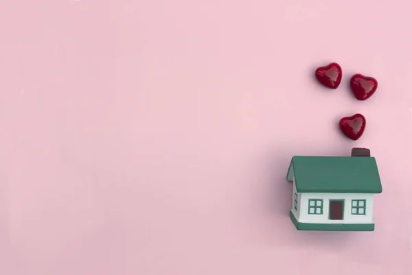Küçük bir ev, pembe arka planda kırmızı kalpler uçuyor.