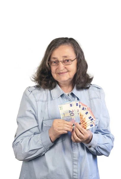 Gözlüklü ve kot gömlekli mutlu yaşlı bir kadın gülümsüyor ve elinde 20 ve 10 avroluk banknotlar tutuyor.