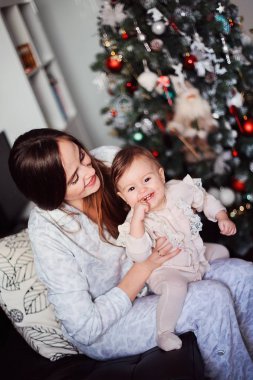 Mutlu güzel kadın Noel ağacının yanında kızıyla eğleniyor. Anne kollarında neşeli bir bebek tutuyor. 