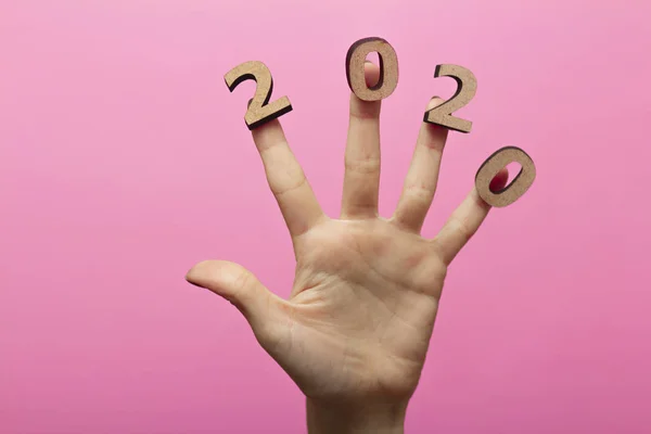Weibliche Hand begrüßt das neue Jahr 2020 auf farbigem rosa Hintergrund, hölzerne Zahlenfiguren auf weiblichen Fingern mit Datum — Stockfoto