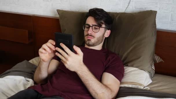 Young man lying in bed and reading ebook — Stock Video