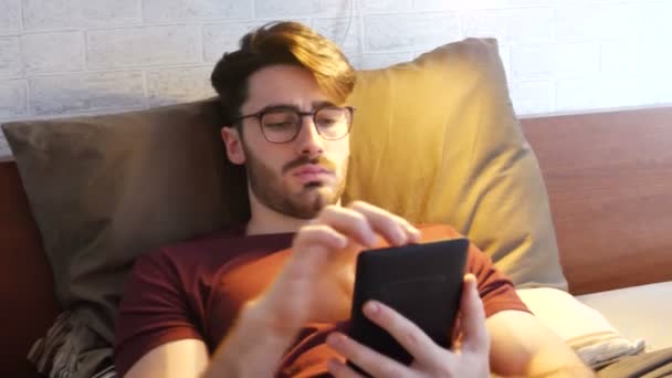 Joven en la cama leyendo con lector de libros electrónicos — Vídeo de stock