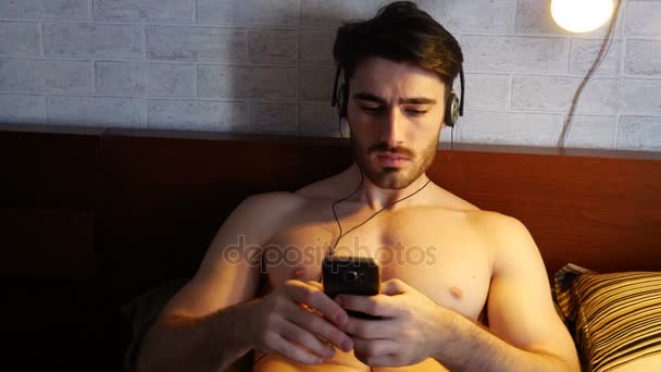 Joven en la cama escuchando música con auriculares — Vídeos de Stock