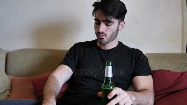 Молодой человек на диване пьет пиво и ест — стоковое видео