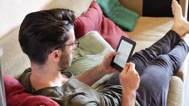 Όμορφος νεαρός άνδρας διαβάζει ebook στον καναπέ — Αρχείο Βίντεο