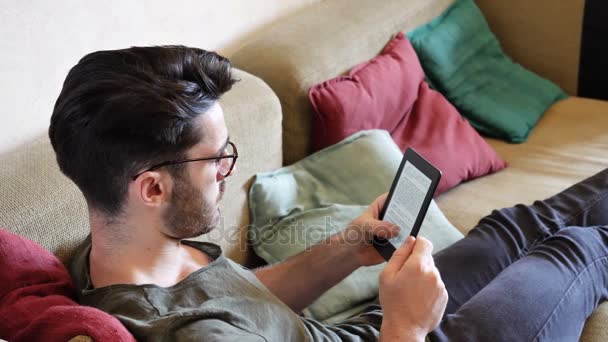 Guapo joven leyendo ebook en sofá — Vídeos de Stock