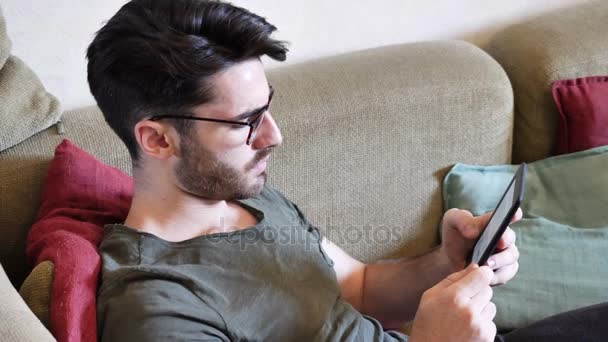 Guapo joven leyendo ebook en sofá — Vídeos de Stock