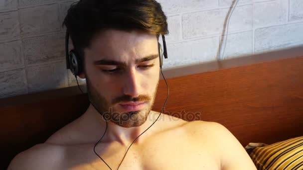 Молодой человек в постели слушает музыку в наушниках — стоковое видео