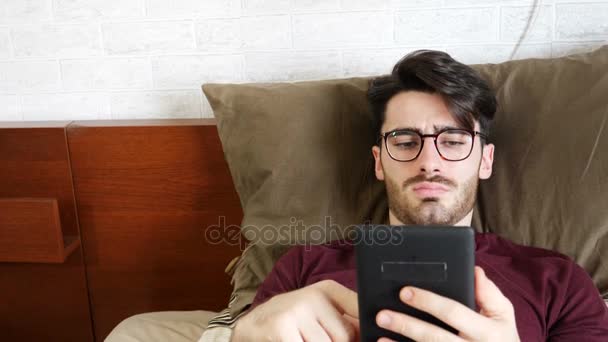 Молодой человек на кровати чтение с электронной книги читателя — стоковое видео
