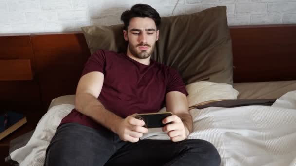 Junger Mann schaut Film auf Handy im Bett — Stockvideo