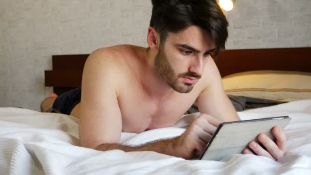 Молодой человек лежит в постели с помощью планшетного компьютера — стоковое видео