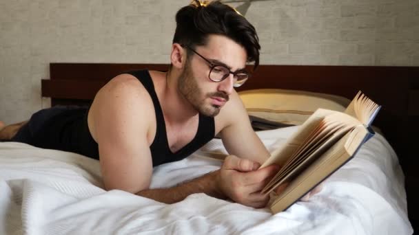 Genç adam yatakta yatan ve bir kitap okuma — Stok video