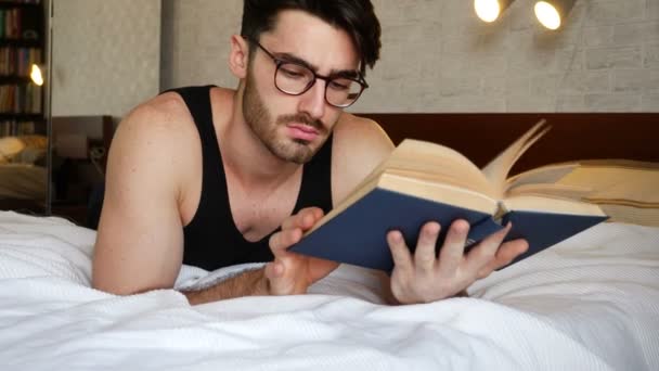 Fiatal férfi feküdt az ágyban, és egy könyvet olvas — Stock videók