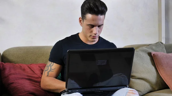 Молодой человек делает домашнюю работу по ноутбуку на диване — стоковое фото