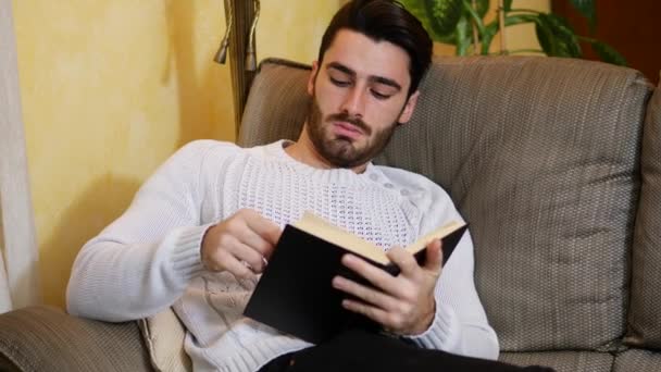 Красивий молодий чоловік читає книгу вдома, сидячи на дивані — стокове відео