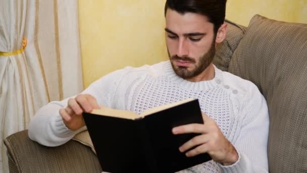 Όμορφος νεαρός άνδρας, διαβάζοντας το βιβλίο στο σπίτι, που κάθεται στον καναπέ — Αρχείο Βίντεο