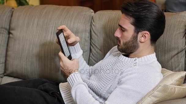 Joven guapo en casa leyendo con lector de libros electrónicos — Vídeo de stock