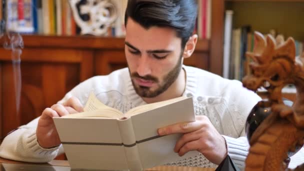 Молодой человек читает книгу дома в своей гостиной — стоковое видео