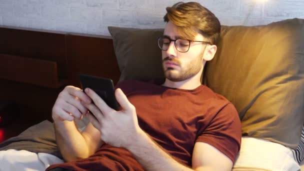 Joven acostado en la cama y leyendo ebook — Vídeos de Stock