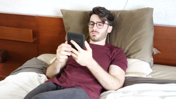 Joven acostado en la cama y leyendo ebook — Vídeo de stock