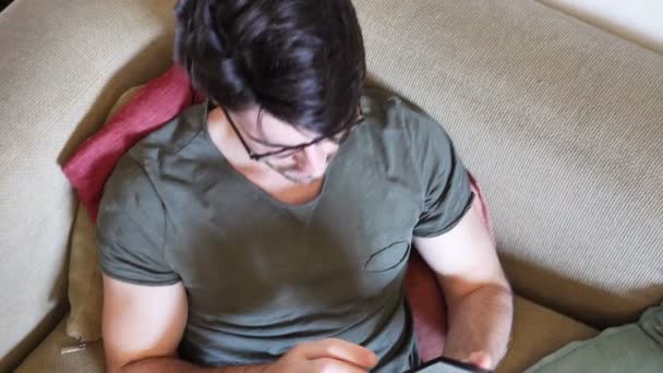 Красивый молодой человек читает электронную книгу на диване — стоковое видео