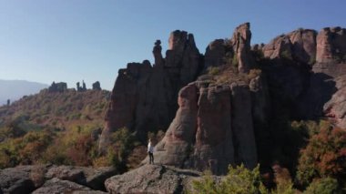 Beyaz gömlekli, özgürlüğünü hisseden genç bir sarışının etrafında uçuşan hava, sonbahar ormanı Belogradchik Kayalıkları, Bulgaristan 'daki çarpıcı kaya oluşumunun kenarında duruyor..