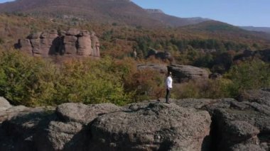 Beyaz gömlekli, özgürlüğünü hisseden genç bir sarışının etrafında uçuşan hava, sonbahar ormanı Belogradchik Kayalıkları, Bulgaristan 'daki çarpıcı kaya oluşumunun kenarında duruyor..