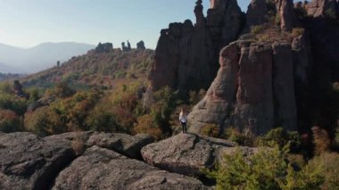 Beyaz gömlekli, özgürlüğünü hisseden genç bir sarışının etrafında uçuşan hava, sonbahar ormanı Belogradchik Kayalıkları, Bulgaristan 'daki çarpıcı kaya oluşumunun kenarında duruyor..