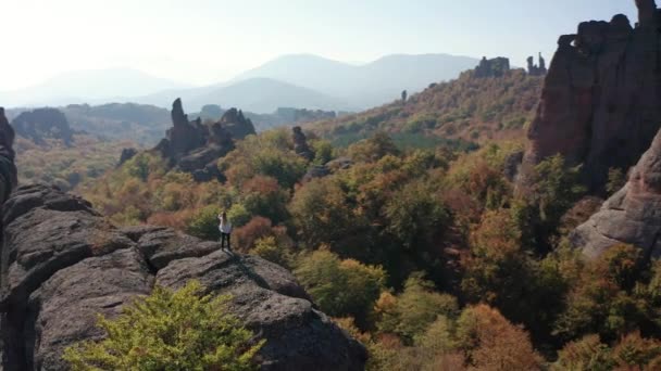 秋の森に囲まれた美しい岩の端に立っている間 白いシャツを着た若いブロンドの女性の周りを空飛ぶ Belogradchik Rocks ブルガリア — ストック動画