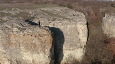 Etrafı sonbahar ormanlarıyla çevrili muhteşem kaya oluşumunun kenarında dikilirken akıllı telefonuyla fotoğraf çeken genç bir kadının etrafında uçan bir hava aracı.