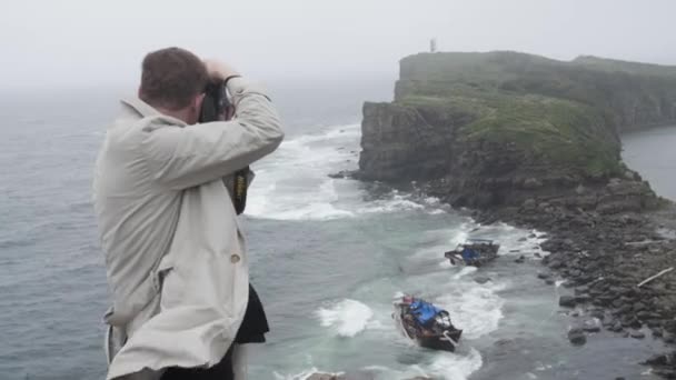 Um fotógrafo de viagem de capa branca tira uma foto de dois barcos azuis quebrados. as consequênciasda tempestade. Tomisina Cape, Ilha Russa, Vladivostok 2019 — Vídeo de Stock