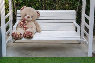Bahçedeki beyaz tahta salıncakta oturan sevimli oyuncak ayı.