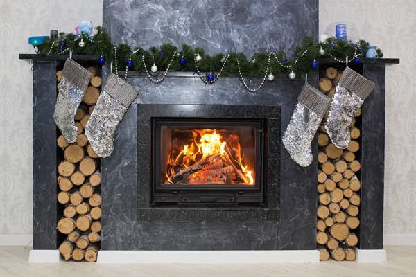 Χριστουγεννιάτικο τζάκι με αναμμένη φωτιά, γιρλάντα και καυσόξυλα για προσάναμμα. Ένα ασημί-γκρι τζάκι στον τοίχο με κάλτσες για χριστουγεννιάτικα δώρα. — Φωτογραφία Αρχείου