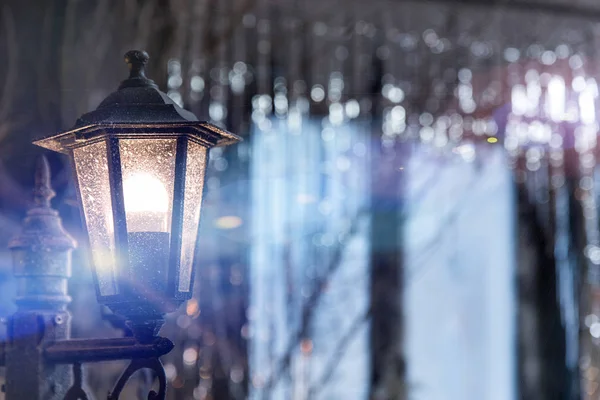 Antique frosty lamppost with blue light on the street close up on the background of the window in shining Christmas decorations sequins. Vintage glass light in night city. — Stock Photo, Image