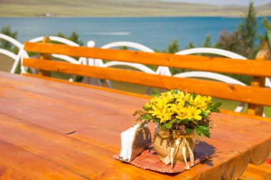 Restoranın terasında üzerinde sarı çiçekler ve peçeteler olan ahşap bir masa. Dağlar, gökyüzü ve su manzaralı.