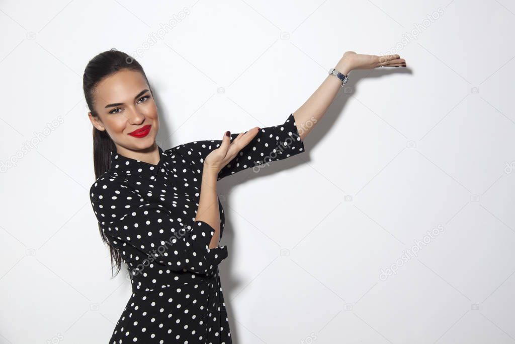Photo of happy young and beutiful woman standing isolated over white wall background. She is looking to camera and showing copyspace pointing. Product placement