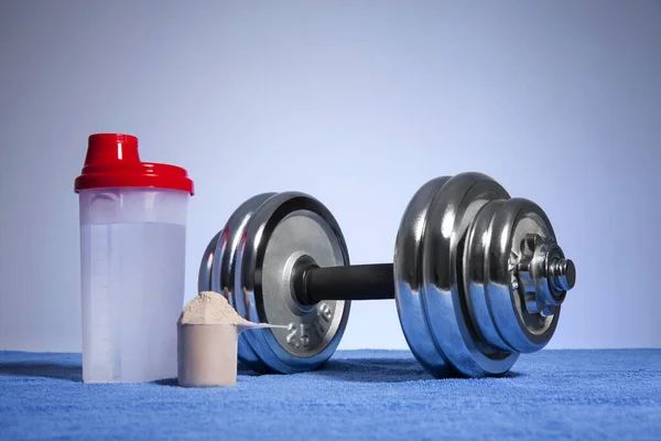 Dumbbell and whey protein shaker on a blue towel surface. Sports bodybuilding supplements or nutrition. Fitness or healthy lifestyle concept.