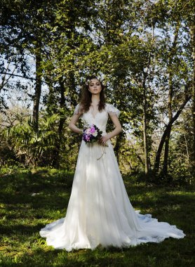 Güzel saçlı, şık bir gelinlik içinde güzel bir gelin ve çiçek buketiyle yeşil arka planda poz veriyor..