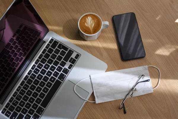 Laptop, cep telefonu, gözlük, kahve ve tek kullanımlık maske çalışma masasında, karantina günlerinde evden çalışıyor. Çalışıyorum, evde kalıyorum. Ders, çalışma masası. Online eğitim. Merkez Ofis.