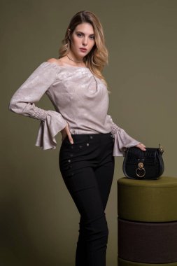 Beyaz bluz, siyah pantolon, yüksek topuklu ayakkabı ve siyah çantayla poz veren zarif sarışın manken. Elini beline koy. Zeytin yeşili arka plan.