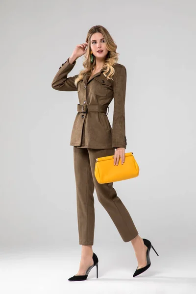 レザーオリーブグリーンのスーツに身を包んだ豪華なブロンドモデル 黄色いハンドバッグ 立って 白地だ スタジオショット — ストック写真