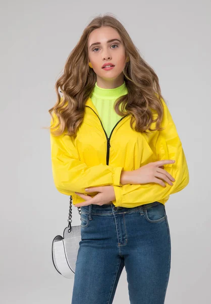 10代のブロンドの女の子が白いワニ革のバッグと青いデニムジーンズで黄色のレインコートに身を包んだ 白い背景のスタジオで — ストック写真