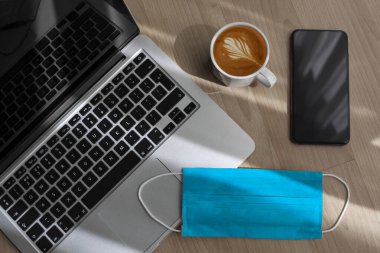 Çalışıyorum, evde kalıyorum. Ders, çalışma masası. Online eğitim. Merkez ofis. Laptop, cep telefonu, kahve ve tek kullanımlık maske çalışma masasında, karantina günlerinde evden çalışıyor.