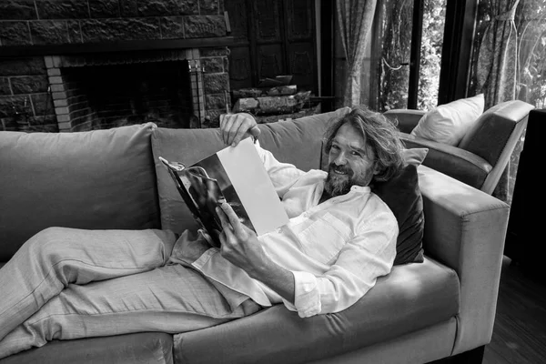 Man in a white shirt. Spending time at home and relaxing. Lying down on the couch, having a good time, reading funny content on the magazine. Looking at camera and laughing, smiling.